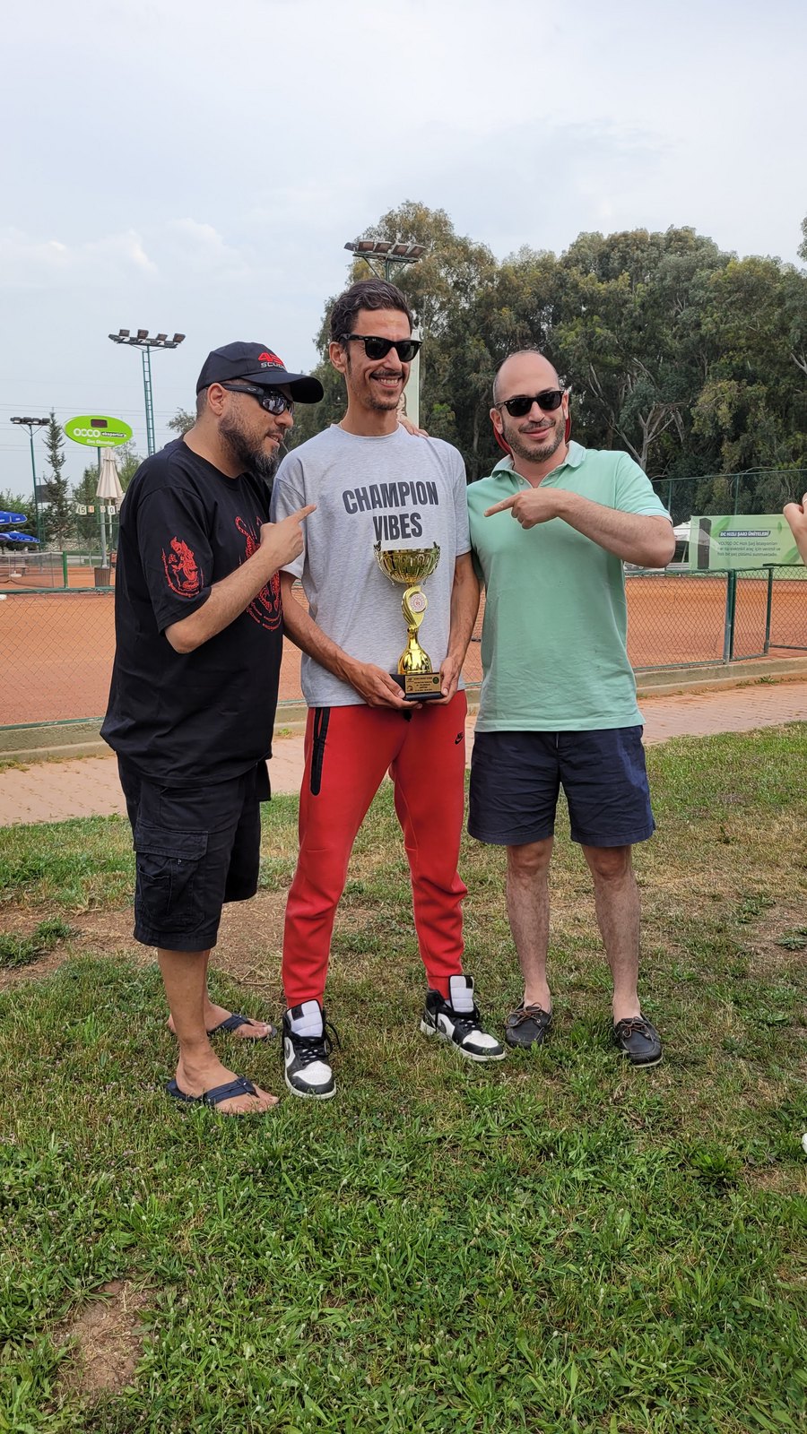 Three friends outdoors at a tennis club, the middle one wearing a grey CHAMPION VIBES tee and holding a gold trophy